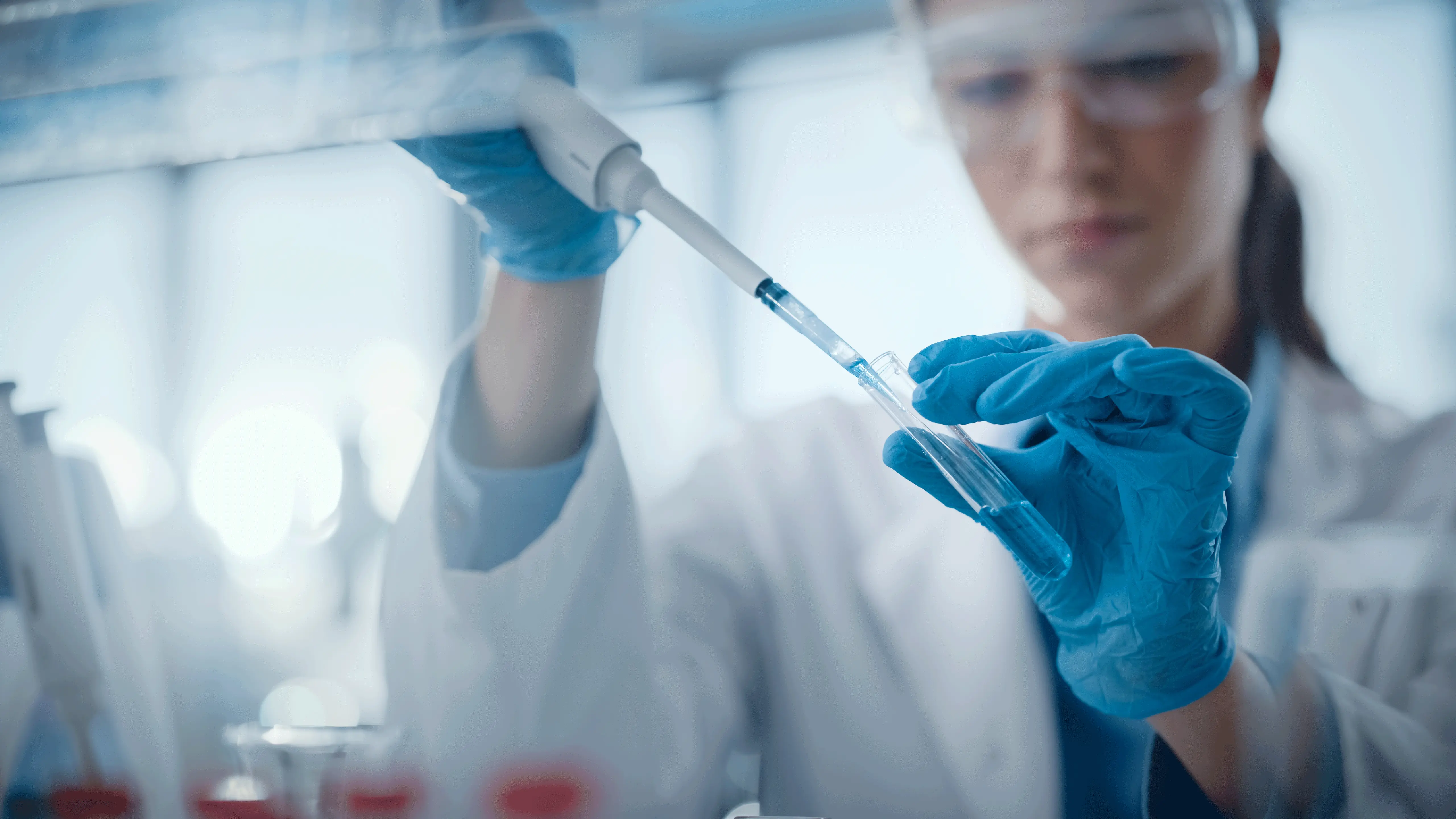 Wissenschaftlerin mit Laborkittel und Schutzausrüstung pipettiert eine Flüssigkeit in ein Reagenzglas in einem Laborumfeld.